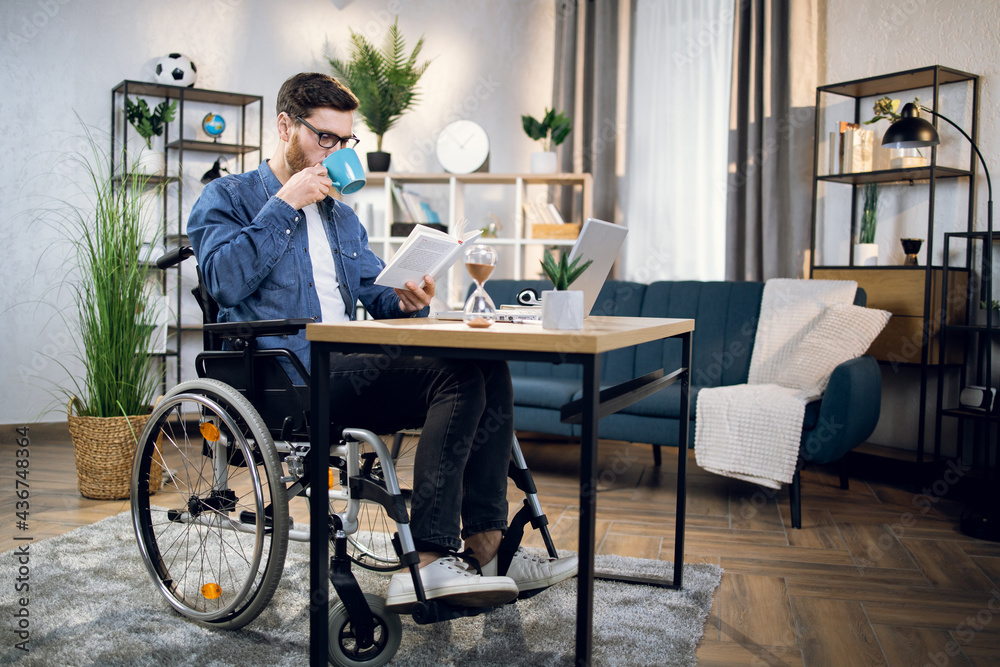 Handicapped young man drinking coffee and reading book while while sitting at table with modern laptop. Male freelancer in wheelchair relaxing during remote work at home.