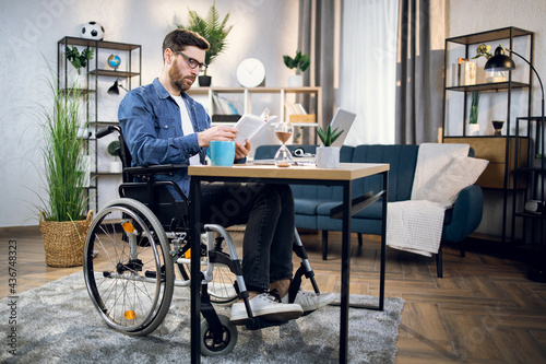 Handicapped young man drinking coffee and reading book while while sitting at table with modern laptop. Male freelancer in wheelchair relaxing during remote work at home.