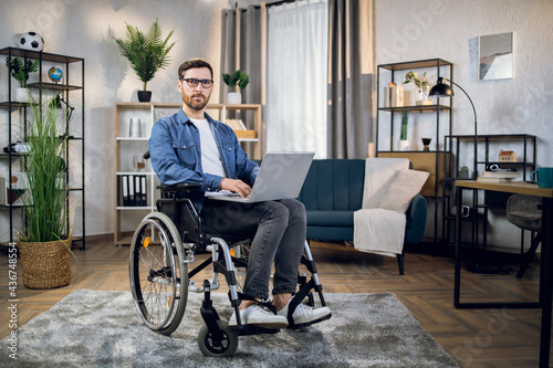 Young male freelancer sitting in wheelchair, using wireless laptop and looking at camera. Remote work for disabled people. Technology concept.