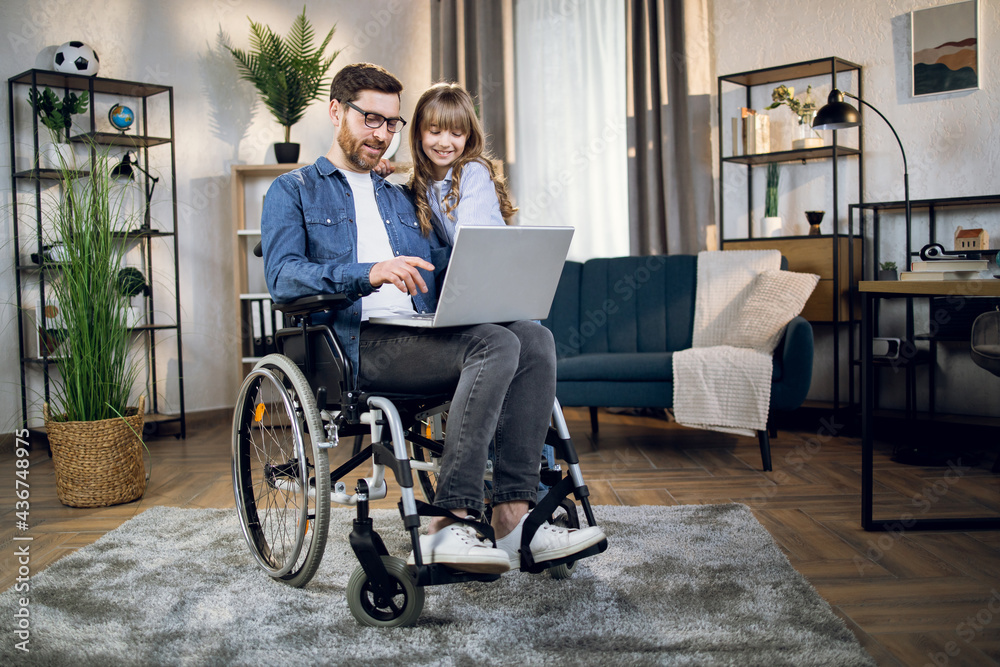 Cute little daughter standing near her father that sitting in wheelchair and working on laptop. Support from beloved family. People with special needs. Cute little daughter standing near her father that sitting in wheelchair and working on laptop. Support from beloved family. People with special needs.