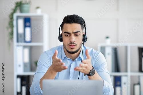 Concerned young Arab man in headset making online video call with business partner, solving problem at office