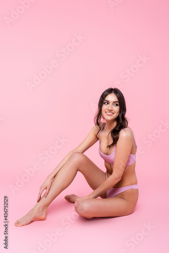 Cheerful brunette woman in swimsuit sitting on pink background.