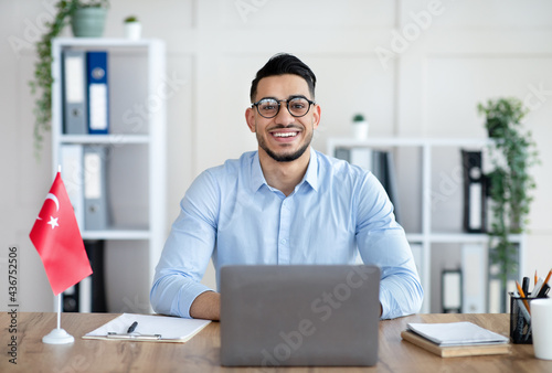 Portrait of millennial male teacher with laptop teaching Turkish online from home, smiling at camera photo