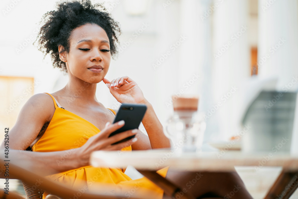 Young woman working smiling typing a message on her phone. African-American appearance and black curly hair. Sitting in a summer restaurant after a pandemic. Online training. Young woman working smiling typing a message on her phone. African-American appearance and black curly hair. Sitting in a summer restaurant after a pandemic. Online training.