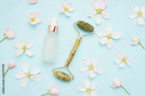 Jade roller for face massage, glass dropper bottle, and apple tree blossom on a blue background. SPA concept