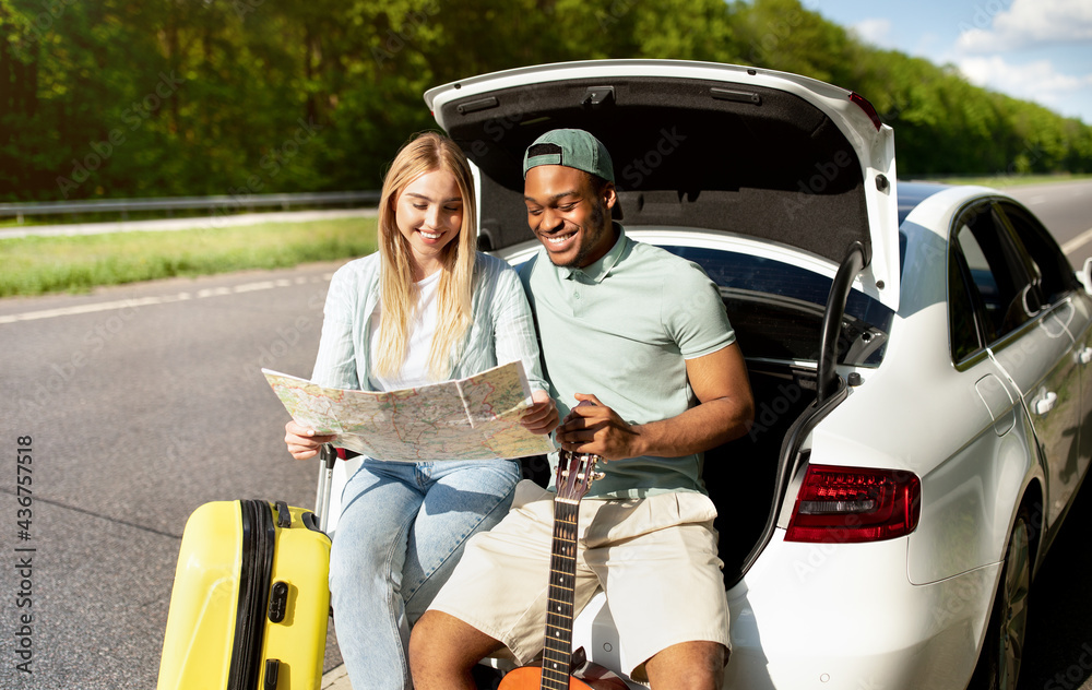 Naklejka premium Multiracial couple going on road trip in car, looking at map, taking break from driving, sitting in auto trunk together