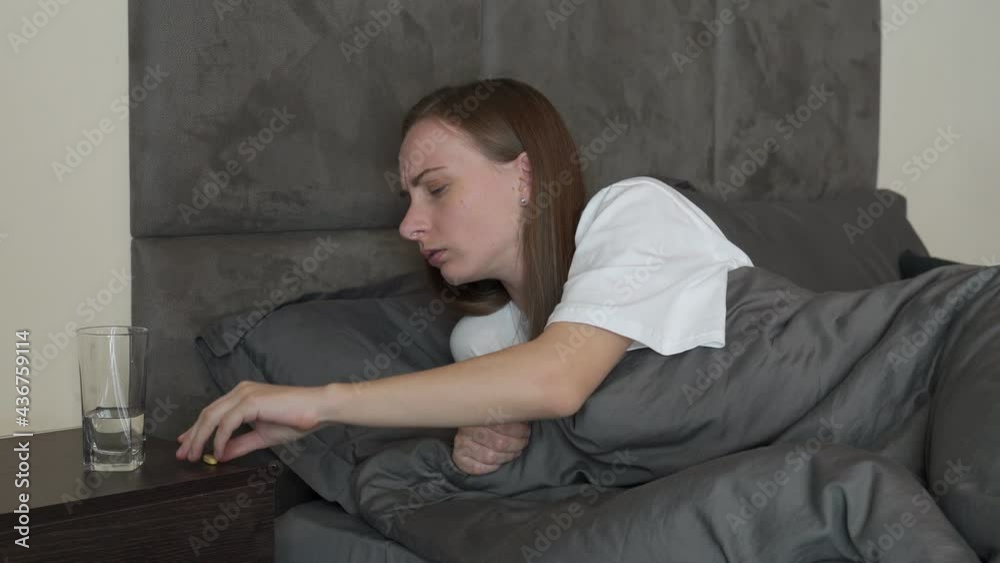 Sad, worried woman is lying on the bed, taking a pill. Concept of suffering from terrible pain Sad, worried woman is lying on the bed, taking a pill. Concept of suffering from terrible pain