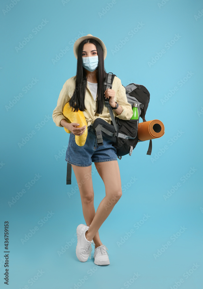 Female tourist in medical mask with backpack and travel pillow on light blue background