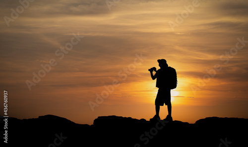 Silhouette of hiker man holding binoculars on sunset from mountain top. Old man with backpack watching the sunset