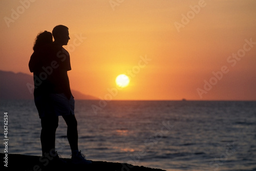 Silhueta de casal apaixonado abraçado e em pé contemplando pôr-do-sol de fronte ao mar. 