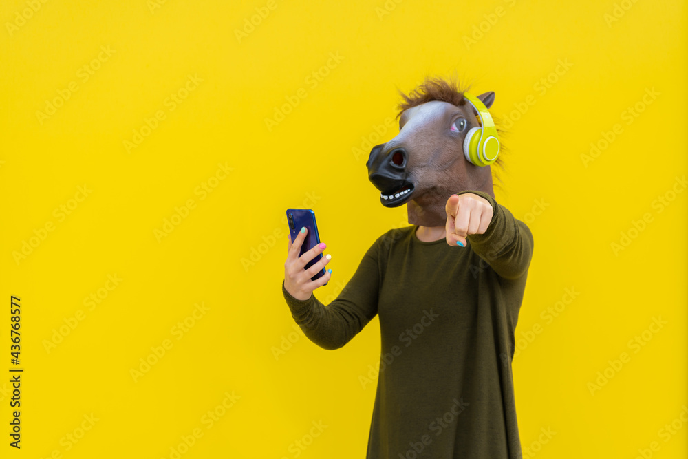 Woman with horse head mask and headphones using smartphone ,pointing finger at camera isolated over yellow background in studio - obrazy, fototapety, plakaty 