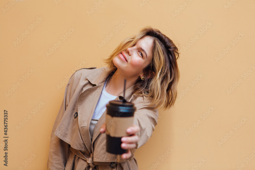 Young smiling millennial woman with wild hair dressed in an autumn coat standing a cup of coffee to go near the beige wall. Young smiling millennial woman with wild hair dressed in an autumn coat standing a cup of coffee to go near the beige wall.