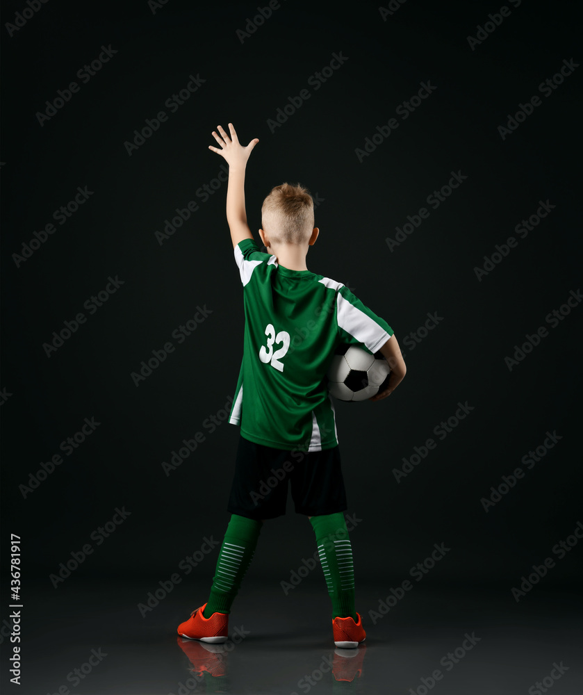 Blond smiling boy child in sports green and white clothes standing holding soccer ball and raised hand up over dark background. Trendy sports active children fashion concept Blond smiling boy child in sports green and white clothes standing holding soccer ball and raised hand up over dark background. Trendy sports active children fashion concept
