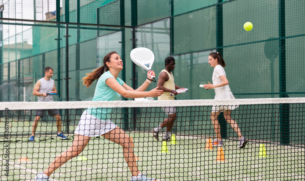 Happy woman learning to play padel game on tennis court outdoor. Other athletes are training in the background