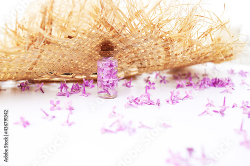 Transparent bottle with lilac flowers standing near a straw hat on a white background