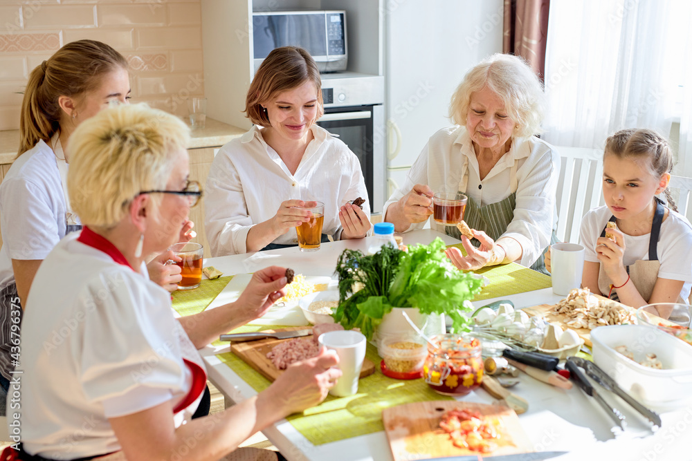 Friendly caucasian family drinking tea and having happy time together, at home in kitchen, have rest after cooking, sitting at table in aprons. family consisted of females of different generations Friendly caucasian family drinking tea and having happy time together, at home in kitchen, have rest after cooking, sitting at table in aprons. family consisted of females of different generations