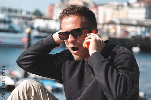 Man with surprised expression talking to his mobile phone at sea port, travel concept