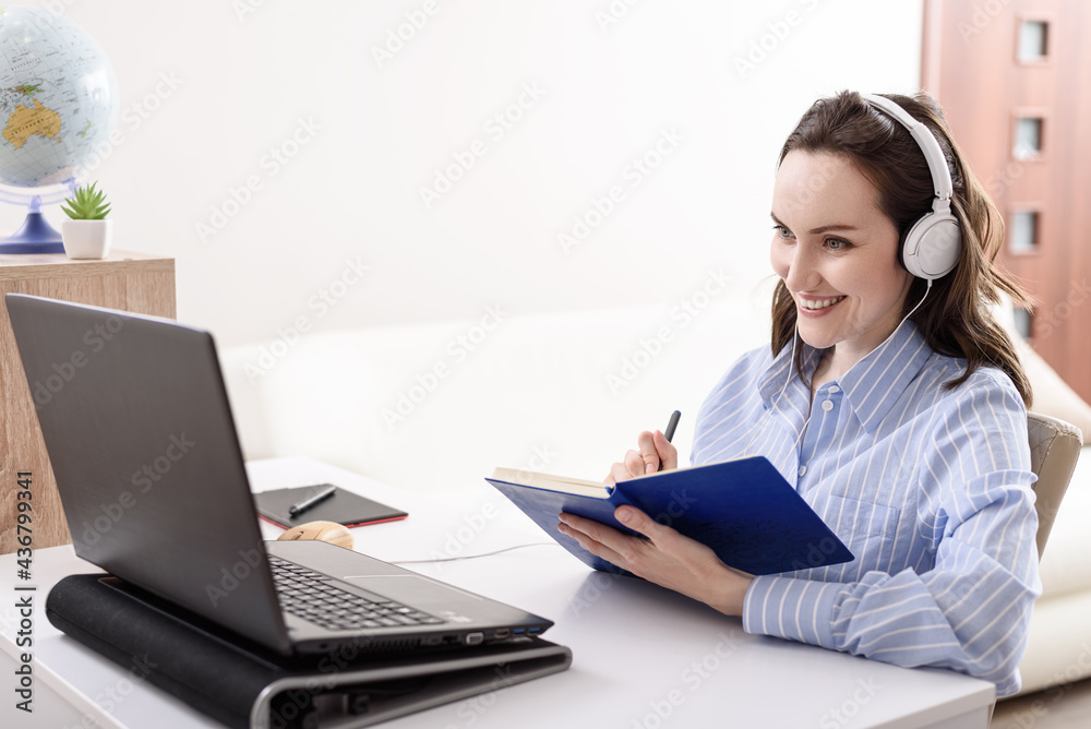 brunette woman in blue shirt with headphones at laptop makes notes in notebook, home-schooled, online courses, remote work at home brunette woman in blue shirt with headphones at laptop makes notes in notebook, home-schooled, online courses, remote work at home