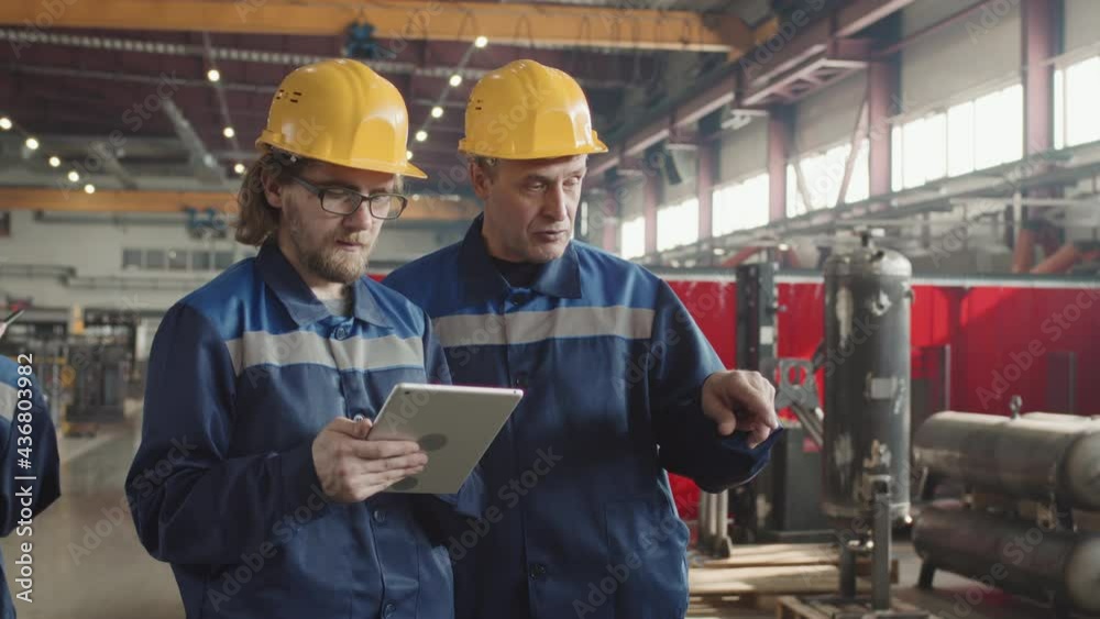 Medium slowmo of senior male supervisor discussing work on plant facility with bearded man holding digital tablet both wearing hard hats and uniforms Medium slowmo of senior male supervisor discussing work on plant facility with bearded man holding digital tablet both wearing hard hats and uniforms