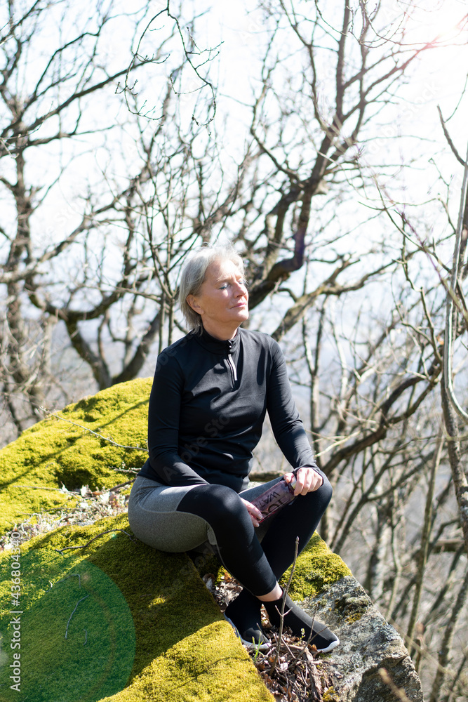 The woman is sitting in a Turkish seat from the profile on a rock covered with moss, in the middle of the forest, her back straight, with her eyes closed in sports equipment. Behind her are bare tree