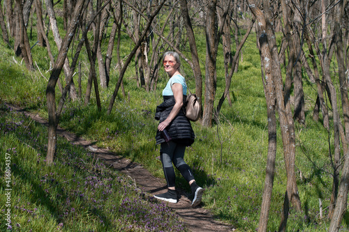 A woman with blonde hair and sunglasses with a bottle of water in her hand, in a sports wardrobe sets through the woods. It is surrounded by bare trees