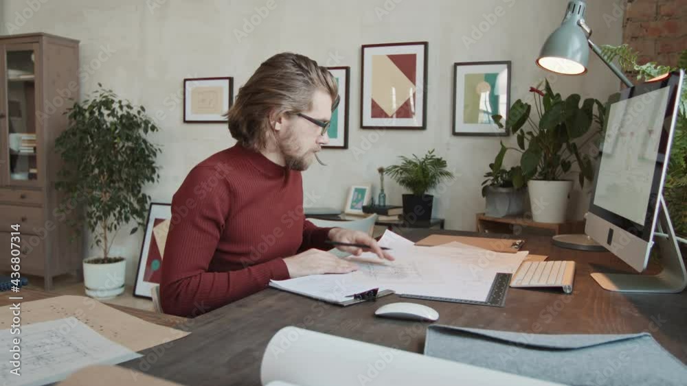 Slowmo portrait of serious male architect sitting at work desk in front of computer and drawing sketches on paper then looking at camera Slowmo portrait of serious male architect sitting at work desk in front of computer and drawing sketches on paper then looking at camera