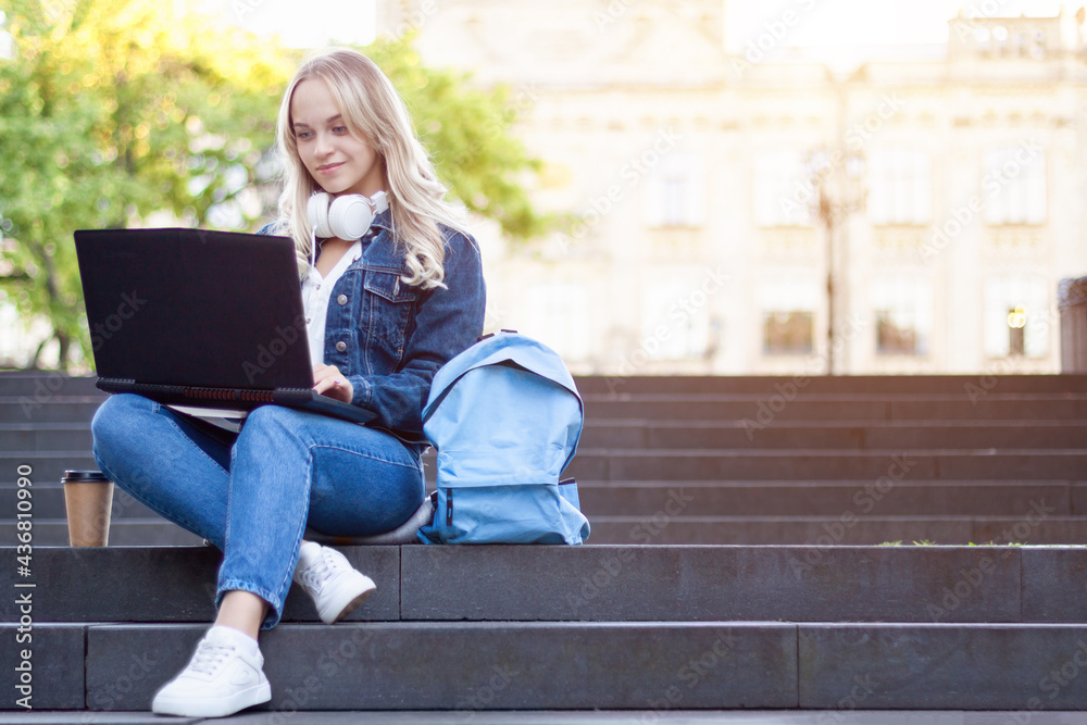 Young blond woman is sitting on stairs, studying with laptop computer, books outdoor near college university building. Girl student is preparing for lecture. High school lifestyle. Education concept.