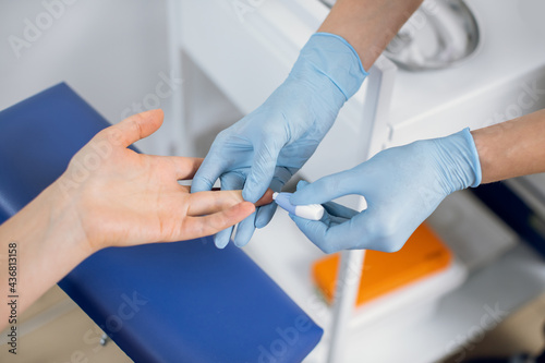 Close up of nurse lab technician in blue sterile gloves, using painless scarifier to prick the finger of unrecognizable patient, sitting on chair. Blood analysis and group determination photo