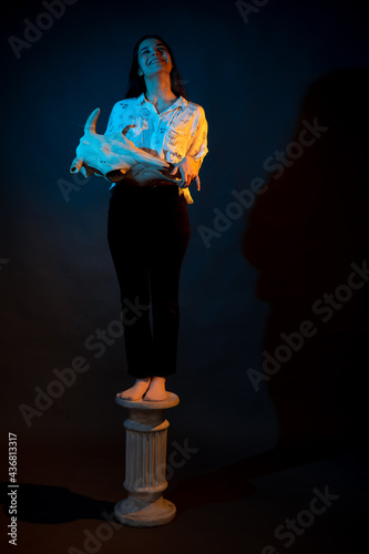 A girl stands on a Greek column with a bull skull in her hands illuminated by blue and yellow light