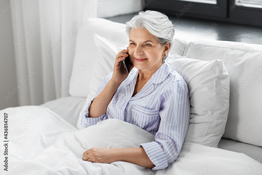 old age, technology and people concept - happy smiling senior woman in pajamas calling on smartphone sitting in bed at home bedroom old age, technology and people concept - happy smiling senior woman in pajamas calling on smartphone sitting in bed at home bedroom