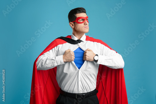 Businessman in superhero cape and mask taking shirt off on light blue background