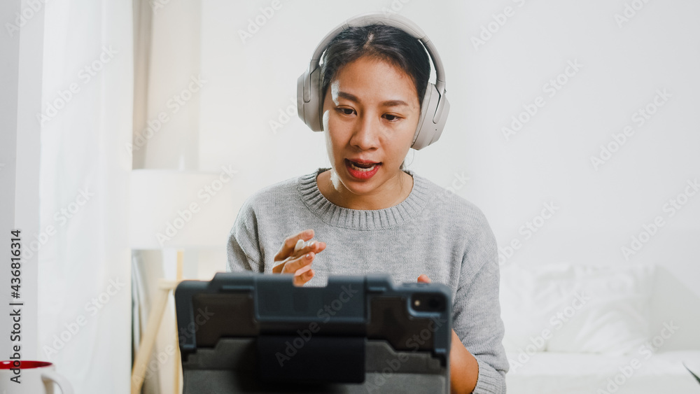 Asia businesswoman wear headphone using tablet talk to colleagues about plan in video call while smart working from home at bedroom. Self-isolation, social distancing, quarantine for corona virus.