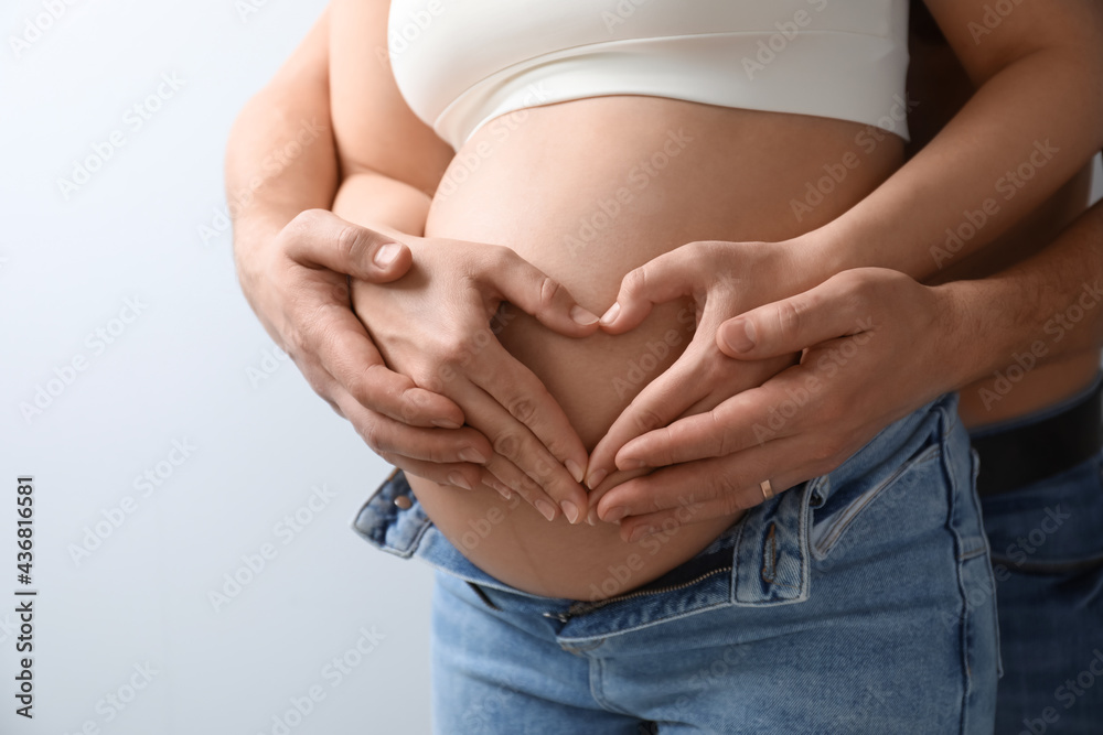 Pregnant young woman making heart with hands on belly and husband near her against light background, closeup Pregnant young woman making heart with hands on belly and husband near her against light background, closeup
