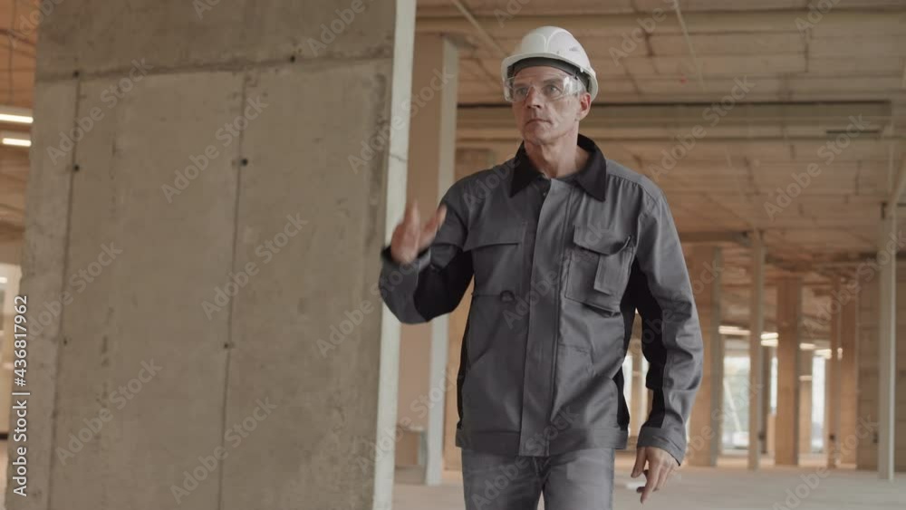 Tracking of middle-aged confident Caucasian male builder wearing worker jacket and white hard hat, walking in building under construction, putting on safety glasses Tracking of middle-aged confident Caucasian male builder wearing worker jacket and white hard hat, walking in building under construction, putting on safety glasses