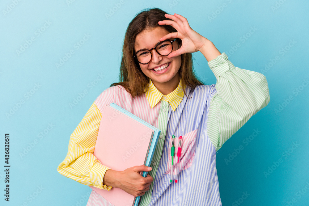 Young caucasian student woman holding books isolated on blue background excited keeping ok gesture on eye. Young caucasian student woman holding books isolated on blue background excited keeping ok gesture on eye.