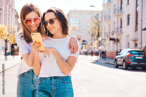 Two young beautiful smiling hipster female in trendy summer white t-shirt clothes and jeans.Sexy carefree women posing on the street background.Positive models using smartphone apps, looking at screen