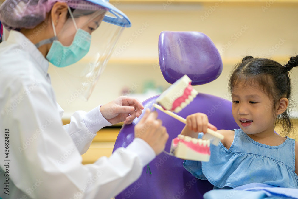 Asian female dentist wearing face mask and face shield teaching a little girl to brush tooth in dental clinic Asian female dentist wearing face mask and face shield teaching a little girl to brush tooth in dental clinic