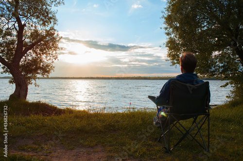Man sitting on the shore of the lake, at sunset. Silhouette of person oncamping chair against backdrop of the setting sun. Outdoor landscape. Fishing and recreation concept.