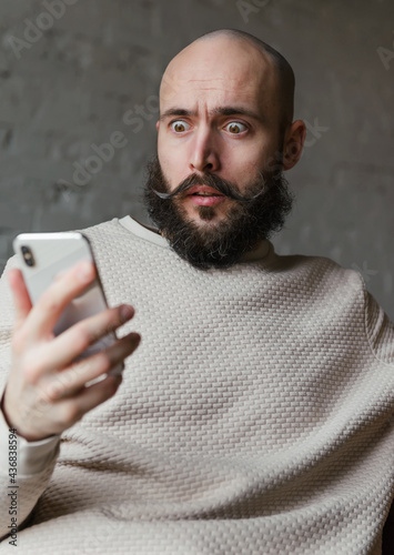 bald man 35 years old with a beard and mustache in a beige jumper looks at the phone with surprise and fear