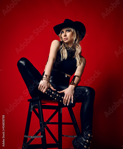Impudent blonde woman in black leather pants, top and stetson hat sits on stepladder holding her feet in hand and looking aside over red background. Fashion, vogue, sexy stylish look for woman concept
