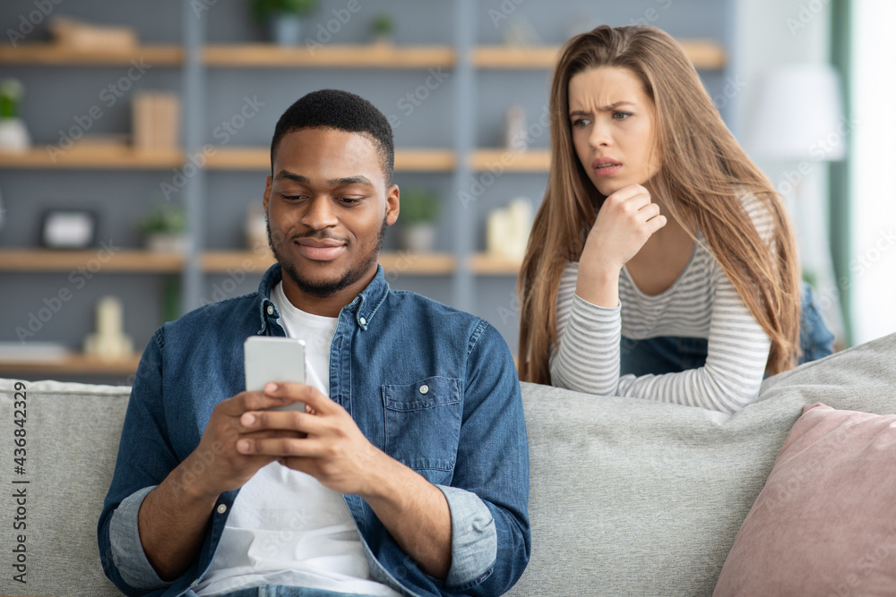 Mistrust. Attentive Lady Looking At Black Boyfriend While He Texting On Smarthone Mistrust. Attentive Lady Looking At Black Boyfriend While He Texting On Smarthone