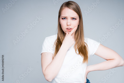 A cute beautiful girl with blond hair in a white T-shirt stands in thought, rubbing her chin with her hand