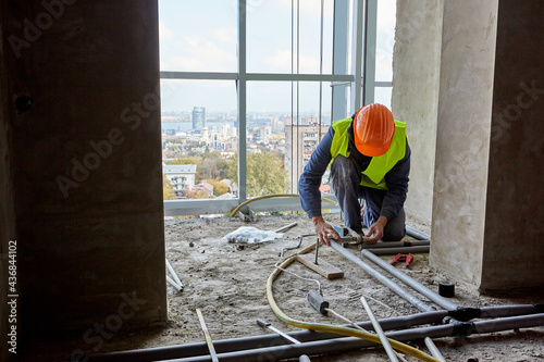 Professional builder in working clothes and safety helmet installing plastic pipes using modern tools in flat of building under construction
