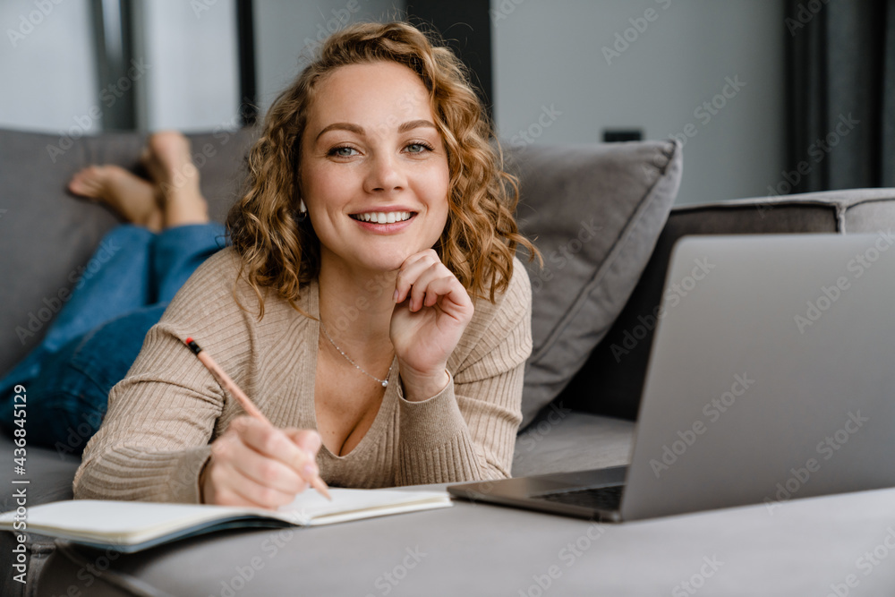 White woman writing down notes and using laptop while lying on couch