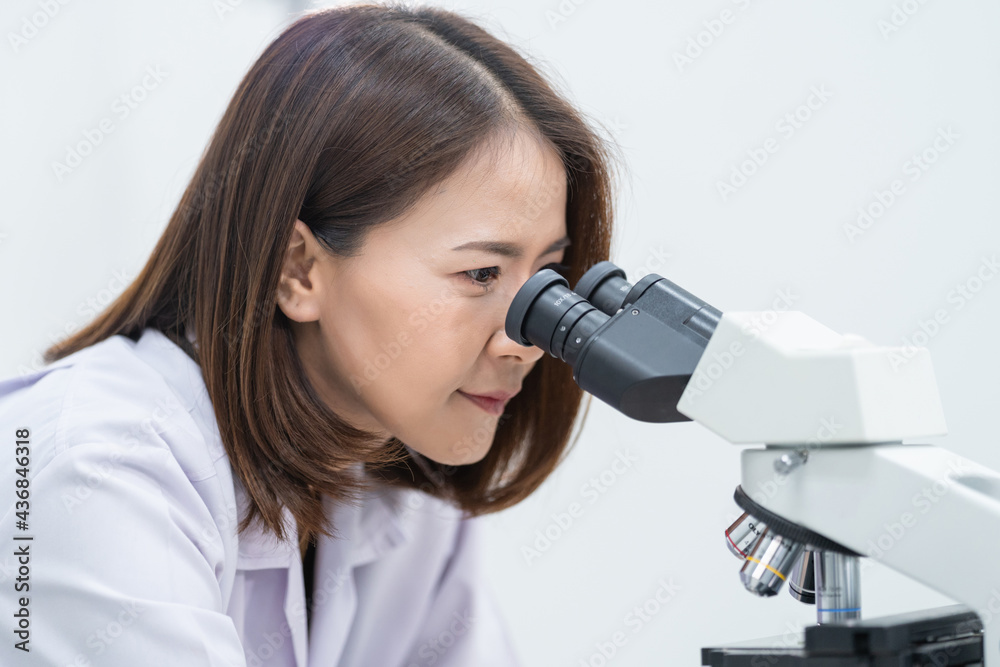 A young scientist woman in a laboratory coat looking through a microscope in a laboratory to do research and experiment. Scientist working in a laboratory. Education stock photo A young scientist woman in a laboratory coat looking through a microscope in a laboratory to do research and experiment. Scientist working in a laboratory. Education stock photo