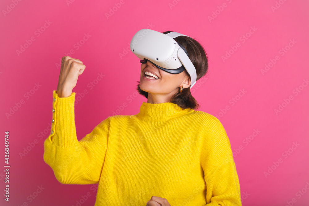 Beautiful woman in bright yellow sweater on pink background in virtual reality glasses happy jumping clenching fist winner gesture Beautiful woman in bright yellow sweater on pink background in virtual reality glasses happy jumping clenching fist winner gesture