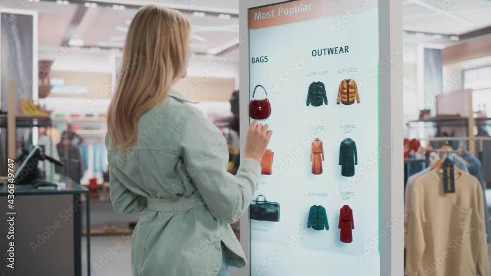 Beautiful Female Customer Using Floor-Standing LCD Touch Display while Shopping in Clothing Store. She is Choosing Stylish Bags, Picking Different Designs from Collection. People in Fashionable Shop.