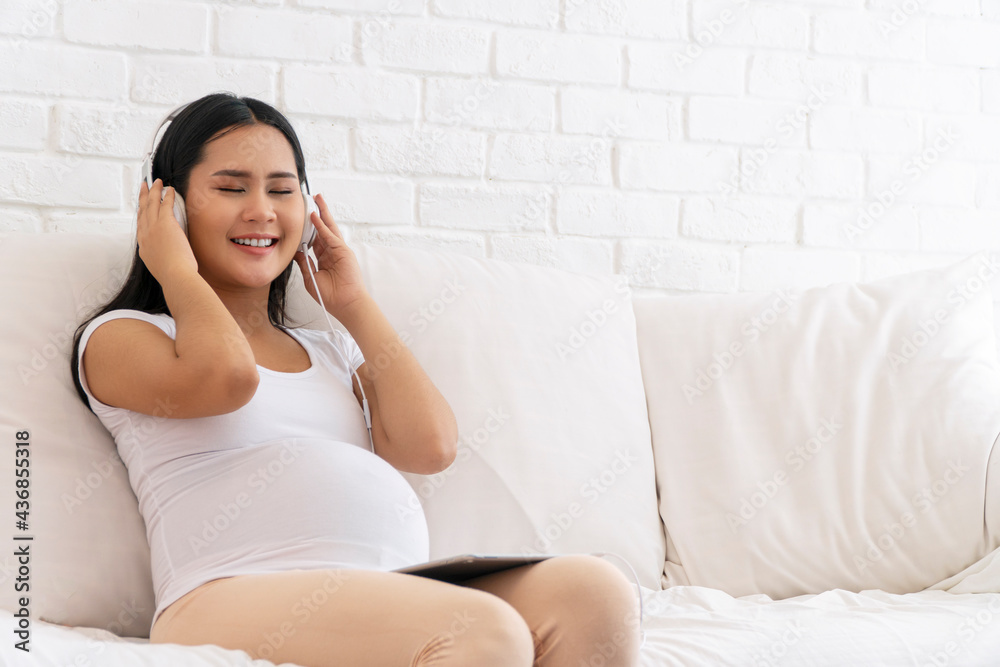 Health care pregnancy concept. Enjoy pregnant young woman wear earphones listening sound music with tablet feeling relaxed while sitting on white sofa at home. Health care pregnancy concept. Enjoy pregnant young woman wear earphones listening sound music with tablet feeling relaxed while sitting on white sofa at home.