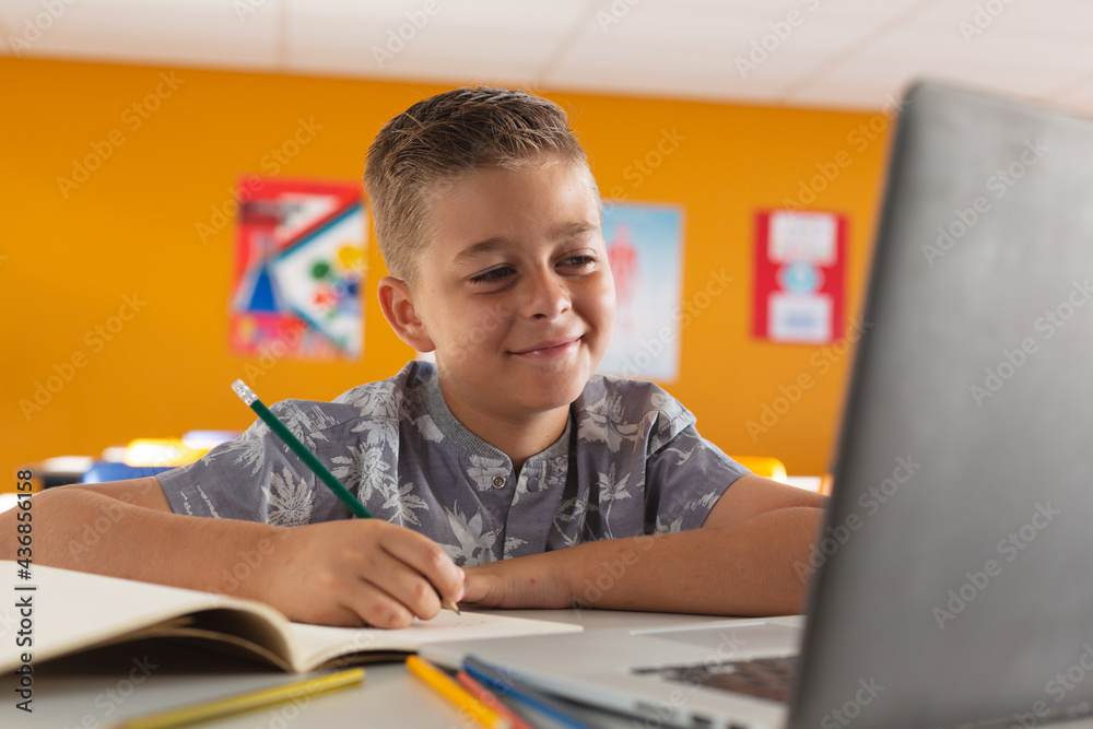 Caucasian boy sitting at a desk in classroom writing, looking at laptop and smiling Caucasian boy sitting at a desk in classroom writing, looking at laptop and smiling