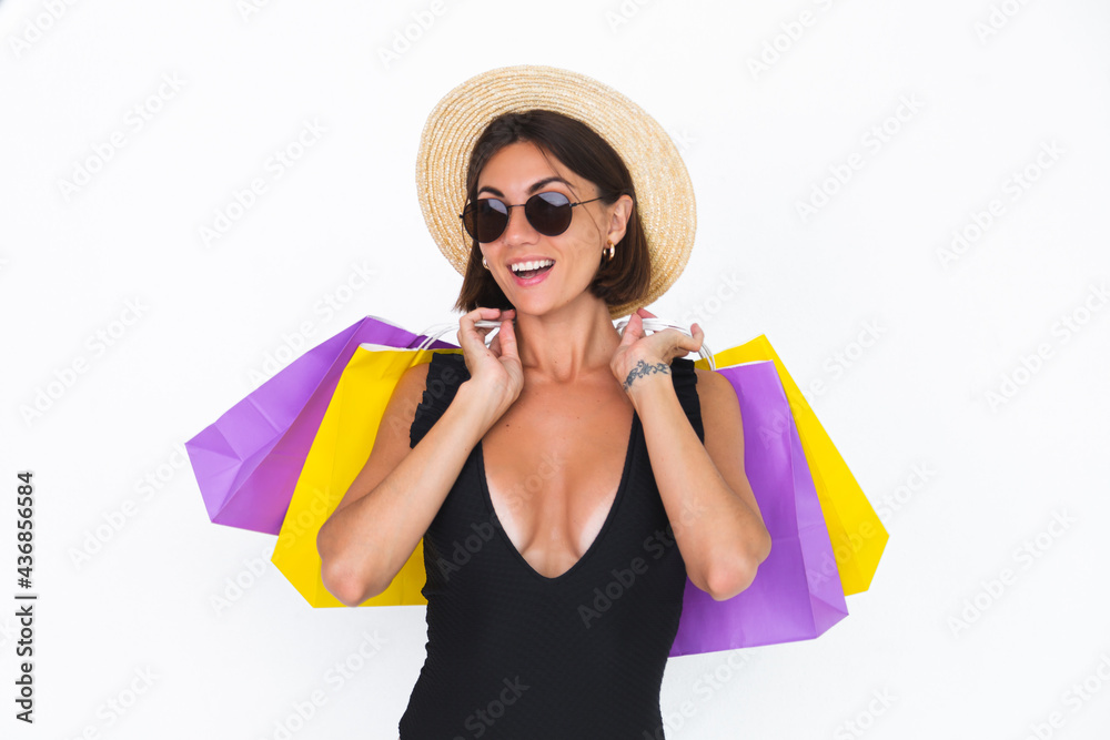 Fit tanned woman in black swimsuit sunglasses and straw hat on white background hold shopping bags happy positive joyful Fit tanned woman in black swimsuit sunglasses and straw hat on white background hold shopping bags happy positive joyful